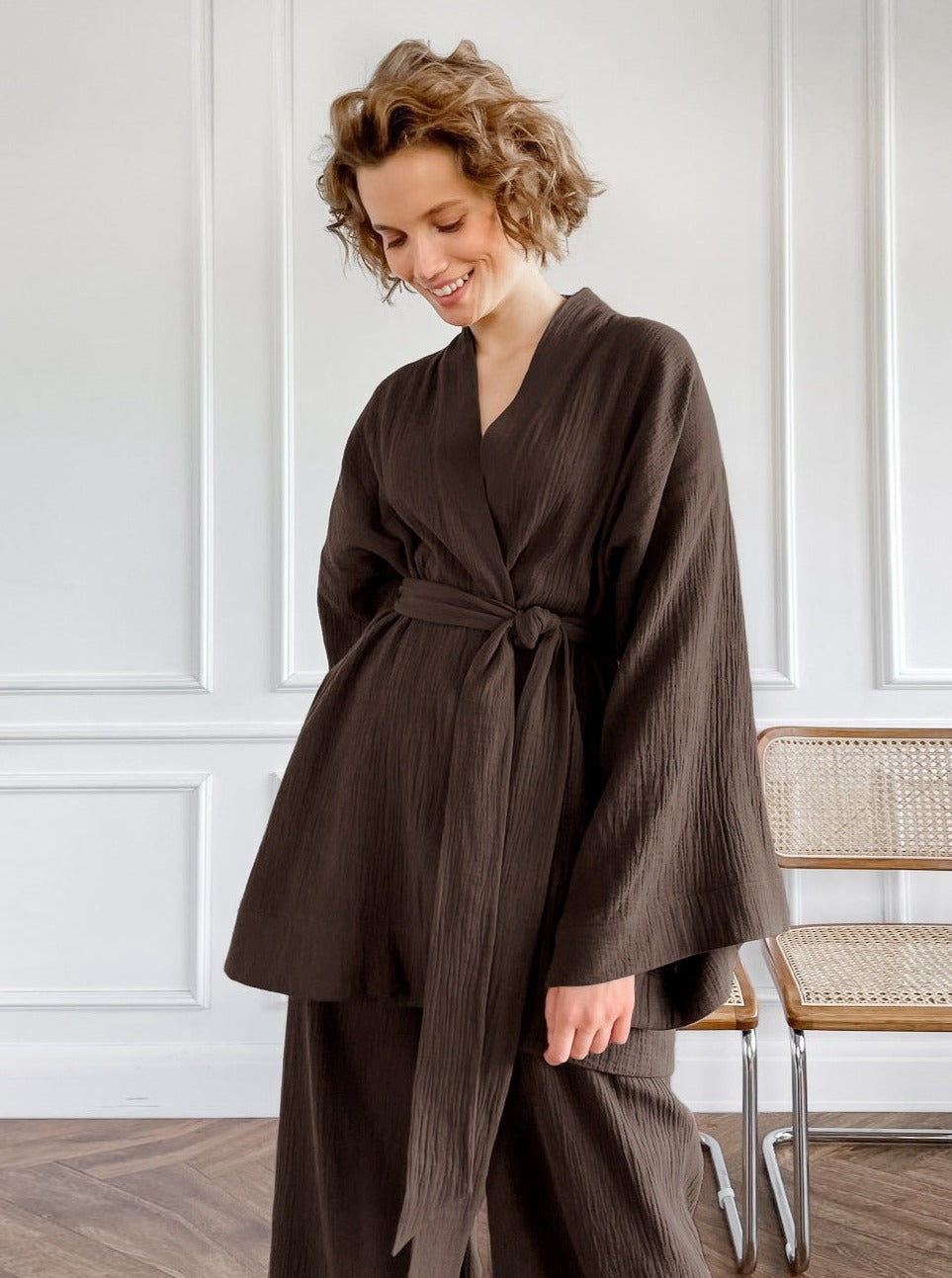 Frau in braunem Kimono-Set, lächelnd, vor weißer Wand. Modetrend, Damenbekleidung, eleganter Look, bequeme Mode, stilvolle Outfits.