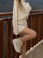Frau in cremefarbenem Strickset und Sneakers posiert auf Balkon mit Bergblick. Modetrend, Strickmode, Herbstoutfit, Damenbekleidung.