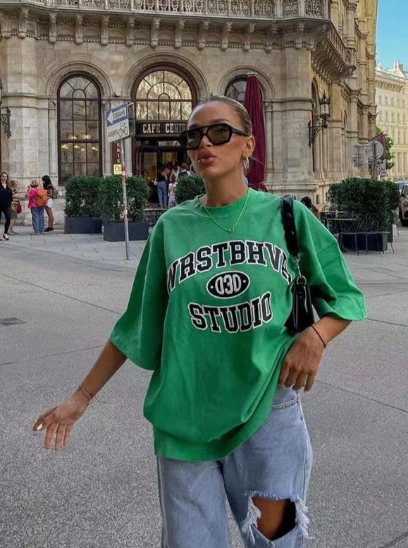 Frau in grünem "Wastbhyve Studio" T-Shirt und zerrissenen Jeans vor Café in urbaner Umgebung, trägt Sonnenbrille und schwarze Tasche.