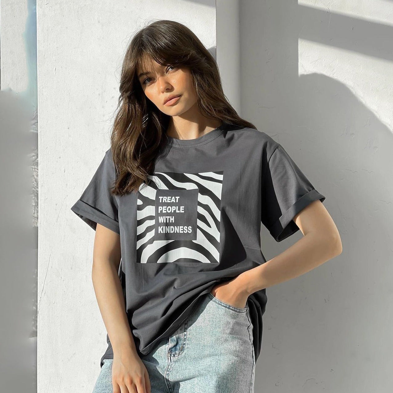 Frau in grauem T-Shirt mit "Treat People With Kindness"-Aufdruck, lässiger Look, modische Damenbekleidung, trendige Streetwear, Jeans.
