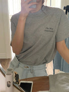 Frau im grauen T-Shirt mit Aufdruck "The Way - Determine", macht Selfie im Spiegel. Modetrend, lässiger Stil, Denim-Jeans, Smartphone.