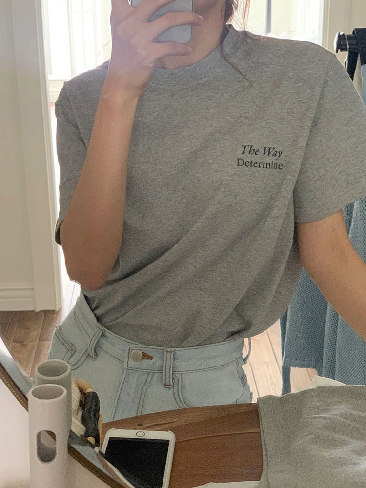 Frau im grauen T-Shirt mit Aufdruck "The Way - Determine", macht Selfie im Spiegel. Modetrend, lässiger Stil, Denim-Jeans, Smartphone.