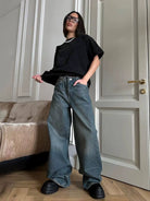 Frau in schwarzem T-Shirt und weiten Jeans posiert in modernem Wohnzimmer. Trendige Mode, lässiger Stil, Oversized-Look, Streetwear.