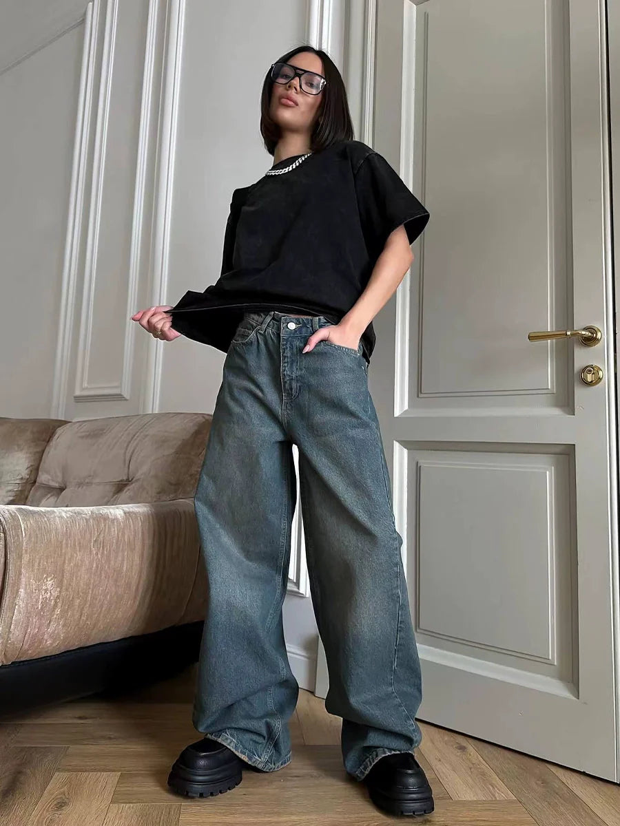 Frau in schwarzem T-Shirt und weiten Jeans posiert in modernem Wohnzimmer. Trendige Mode, lässiger Stil, Oversized-Look, Streetwear.