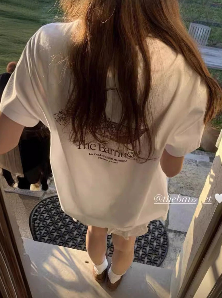 Frau in weißem T-Shirt mit "The Barn" Aufdruck, steht in der Sonne. Mode, Sommer, Freizeitkleidung, Rückansicht, lässiger Stil.