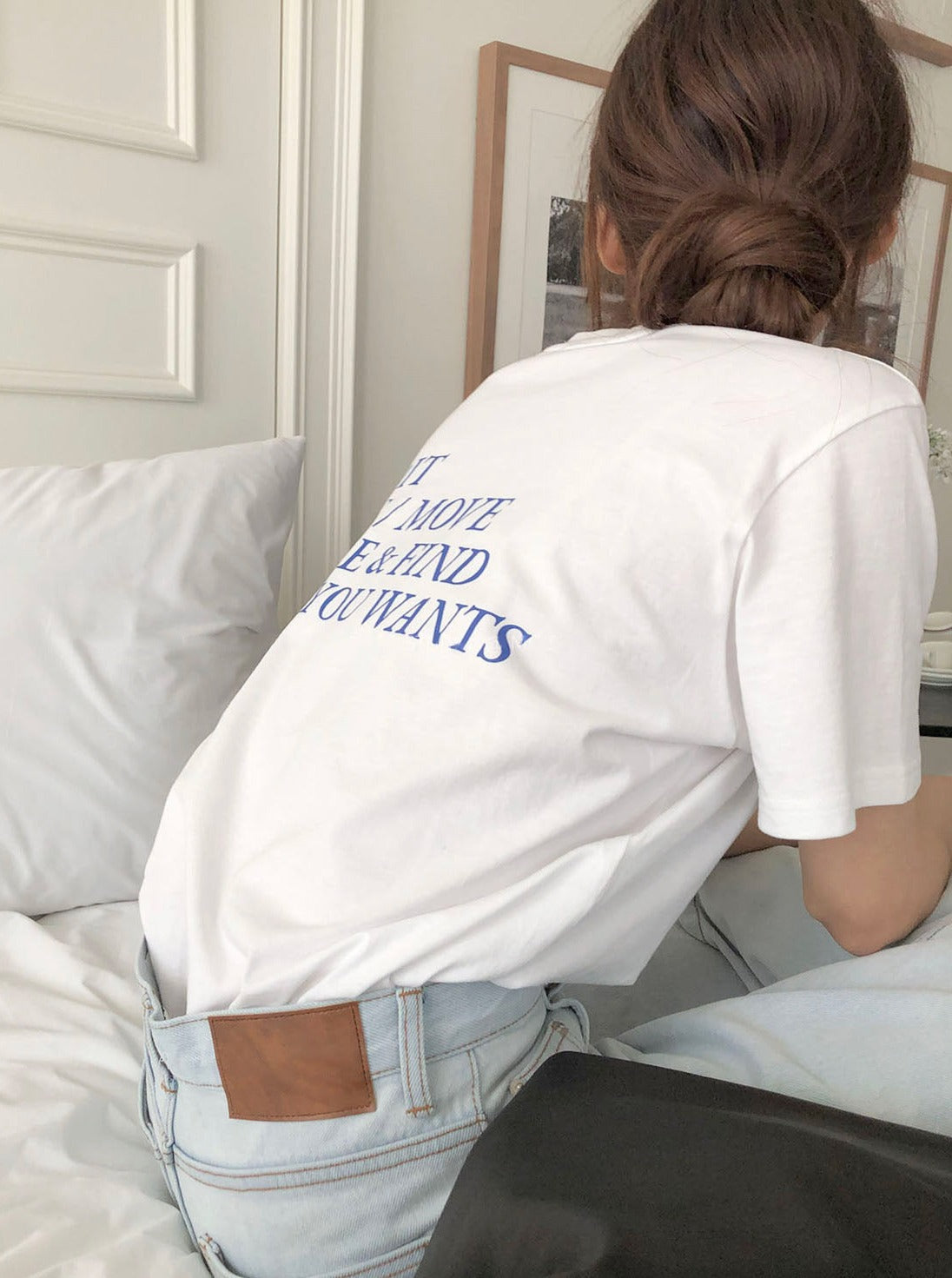 Frau in weißem T-Shirt mit blauer Aufschrift, sitzt auf einem Bett. Modetrend, lässiger Stil, bequeme Kleidung, Freizeitlook, Damenmode.