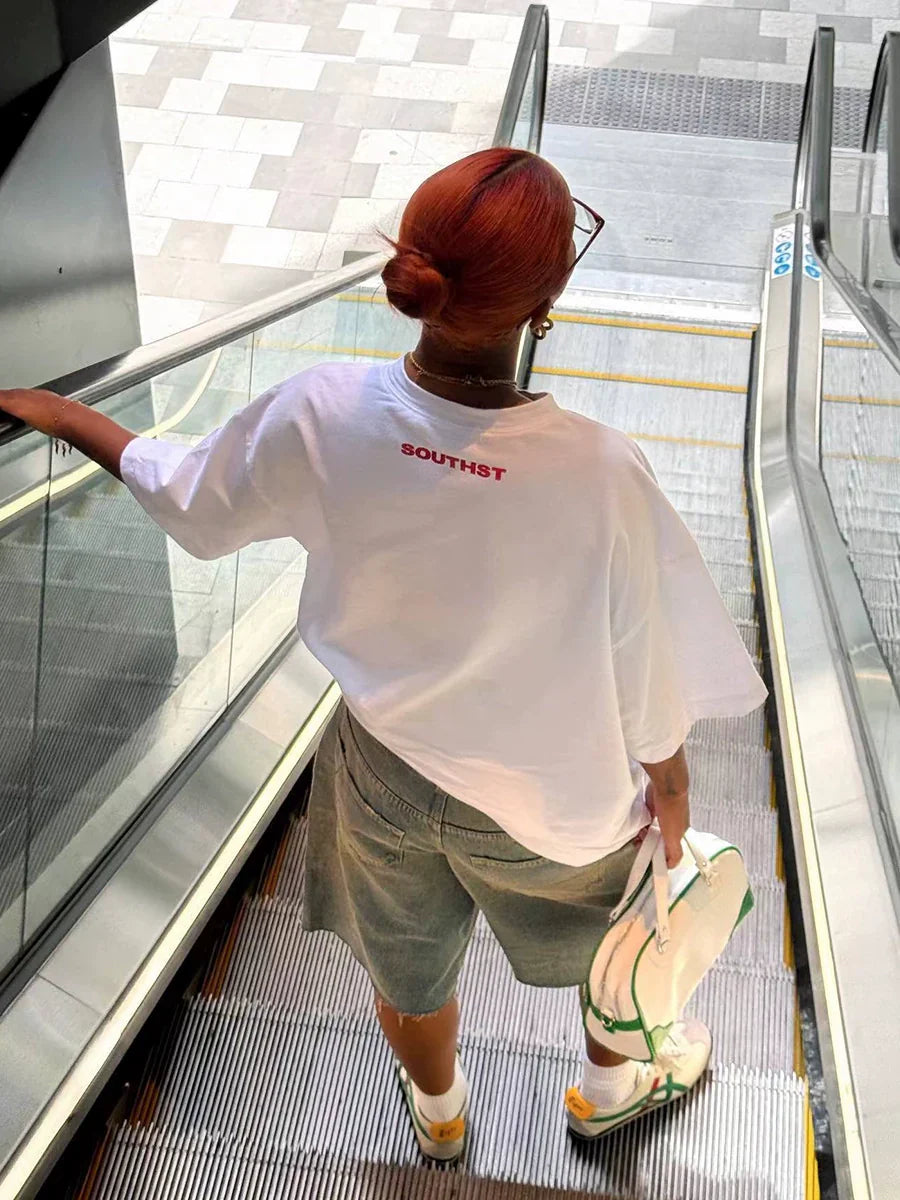 Frau mit rotem Haar auf Rolltreppe, trägt weißes T-Shirt mit "SOUTHST"-Aufdruck, graue Shorts und weiße Sneakers. Urbaner Streetwear-Stil.