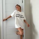 Frau in weißem T-Shirt mit Aufdruck, lehnt an Tür. Modetrend, lässiger Stil, Oversize-Shirt, minimalistische Einrichtung, moderne Kleidung.