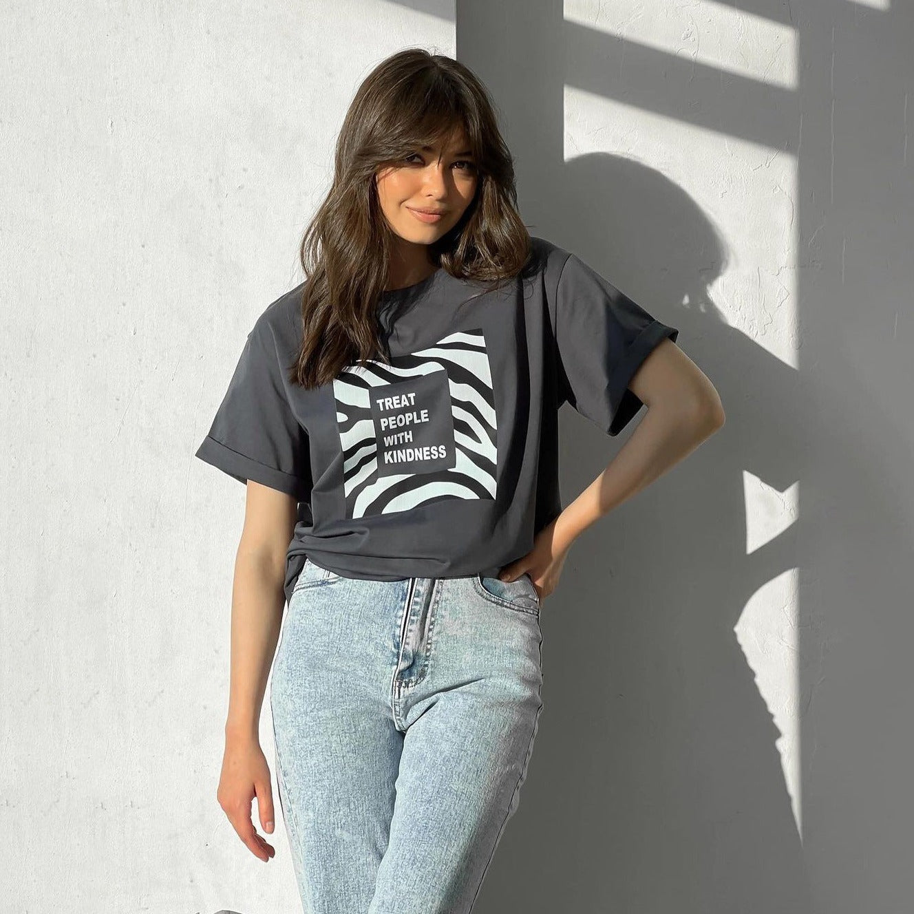 Frau in schwarzem T-Shirt mit "Treat People With Kindness"-Aufdruck, lässig gestylt, kombiniert mit hellblauen Jeans, vor weißer Wand.