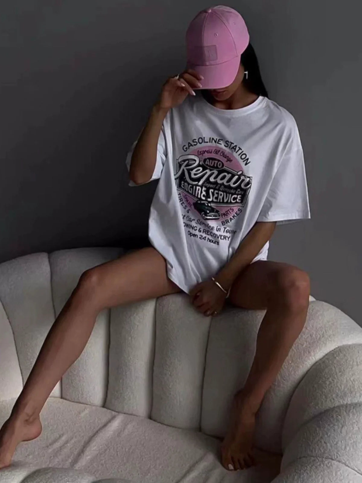 Frau in übergroßem T-Shirt mit "Auto Repair"-Aufdruck, sitzt auf einem weißen Sofa, trägt rosa Baseballkappe. Trendige Freizeitmode, urbaner Stil.