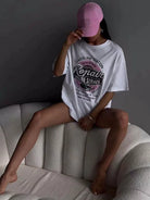 Frau in übergroßem T-Shirt mit "Auto Repair"-Aufdruck, sitzt auf einem weißen Sofa, trägt rosa Baseballkappe. Trendige Freizeitmode, urbaner Stil.