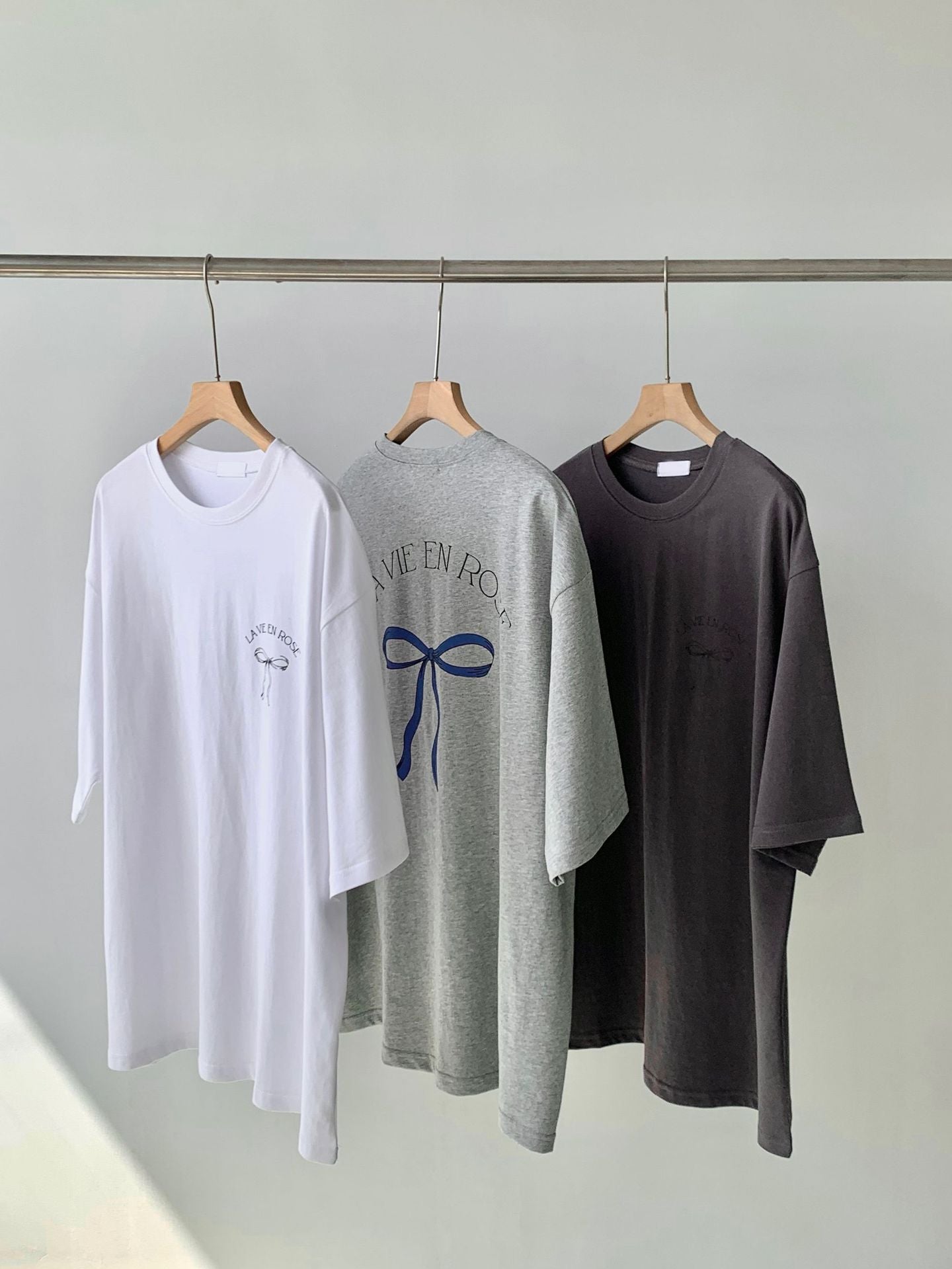 Drei T-Shirts auf Kleiderbügeln: weiß, grau mit Aufdruck "Live en Rose", schwarz. Modetrend, lässige Kleidung, minimalistisch, unisex.