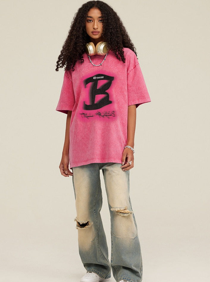 Frau in rosa Oversize-T-Shirt mit schwarzem "B"-Logo, zerrissene Jeans, goldene Kopfhörer; Streetwear, urbaner Stil, modisch, lässig.