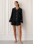 Frau in schwarzem Pyjama-Set mit weißen Paspeln, stehend vor einer eleganten Wand. Nachtwäsche, Damenmode, bequeme Schlafkleidung.