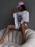 Frau in übergroßem T-Shirt mit Auto-Reparatur-Design, sitzt auf modernem Sofa, trägt rosa Baseballkappe. Trendige Freizeitmode, urbaner Stil.