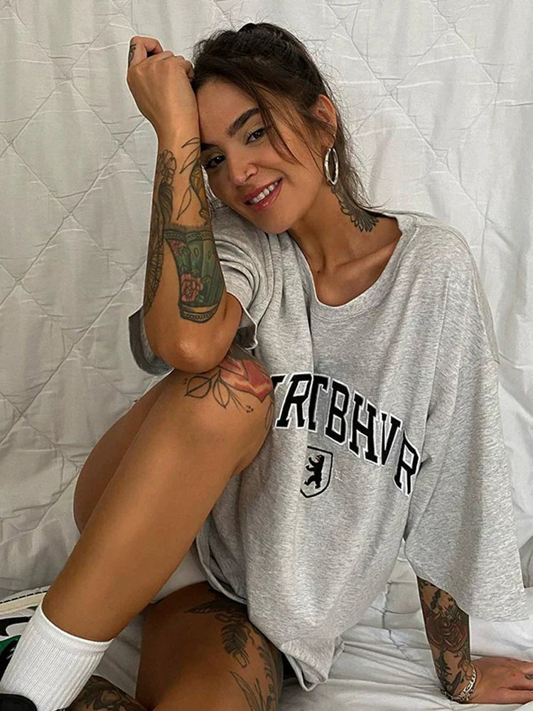 Frau in grauem Oversize-T-Shirt mit "BERLIN"-Aufdruck, tätowierte Arme und Beine, sitzt lächelnd vor weißem Hintergrund. Urbaner Streetwear-Look.