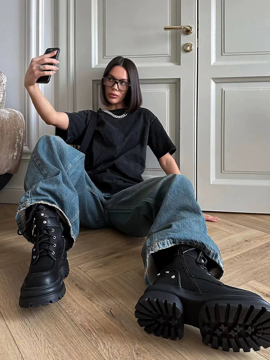 Frau in schwarzem T-Shirt und blauer Jeans macht Selfie, trägt schwarze Stiefel. Trendige Mode, lässiger Stil, Indoor-Fotografie.