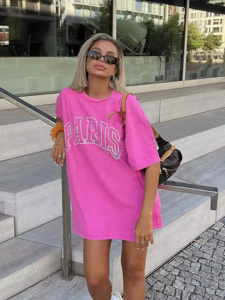 Frau in pinkem Oversize-T-Shirt mit "Paris"-Aufdruck, Sonnenbrille und schwarzem Rucksack. Modetrend, Streetstyle, lässiger Look.