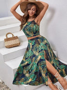 Frau in tropischem Maxikleid mit Blattmuster, sitzt auf Treppe. Sommermode, Strandkleid, Damenbekleidung, Strohhut, Boho-Stil, trendig.