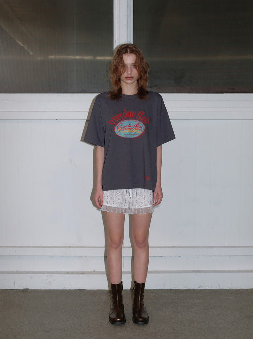 Frau trägt oversized graues T-Shirt mit rotem Print, weiße Spitzen-Shorts und schwarze Stiefel. Modetrend, Streetwear, lässiger Look.