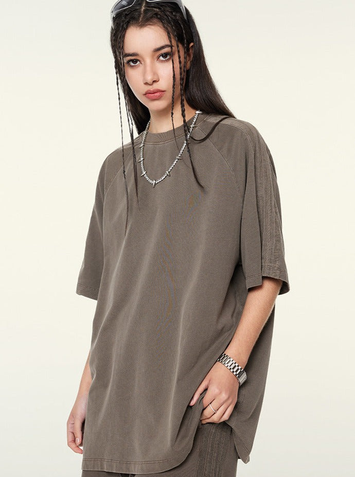 Frau in oversized braunem T-Shirt, lässiger Streetwear-Look, lange Zöpfe, silberne Halskette, modische Sonnenbrille, urbaner Stil.