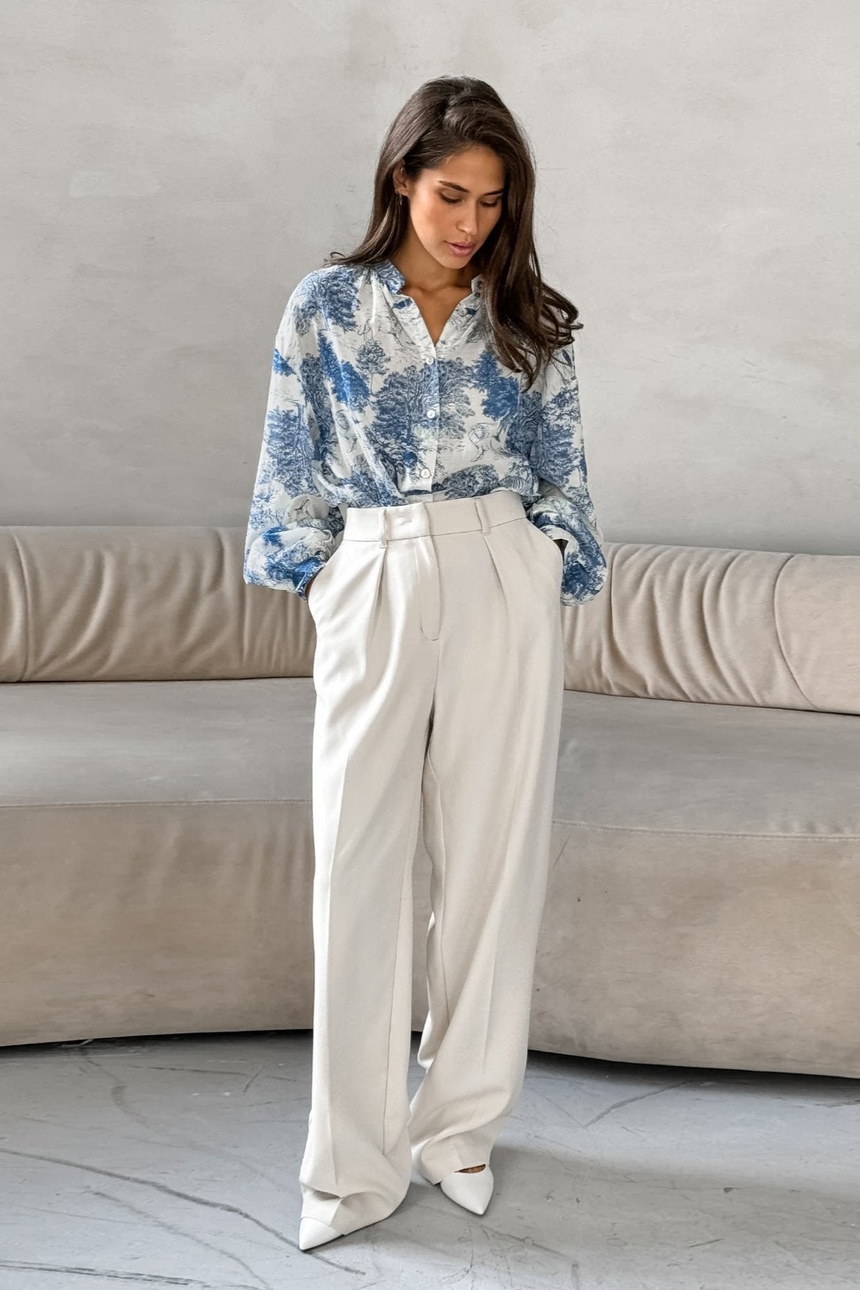 Elegante Frau in weißer Hose und blauer Bluse mit Blumenmuster, steht vor einem beigen Sofa. Modetrends, Damenmode, stilvolle Outfits.