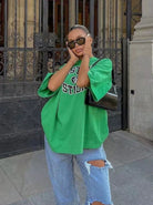 Frau in grünem Oversize-T-Shirt und zerrissenen Jeans vor einem Tor, trägt Sonnenbrille und schwarze Handtasche. Streetwear, lässiger Look.