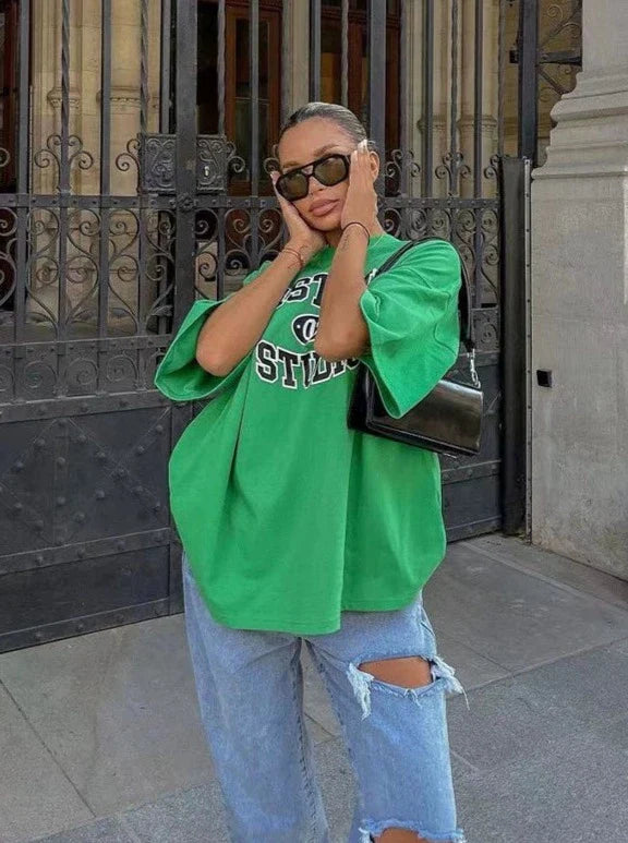 Frau in grünem Oversize-T-Shirt und zerrissenen Jeans vor einem Tor, trägt Sonnenbrille und schwarze Handtasche. Streetwear, lässiger Look.