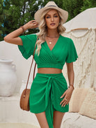 Frau in grünem Sommerkleid mit Strohhut und Umhängetasche, vor mediterranem Hintergrund. Mode, Sommeroutfit, Damenbekleidung, stilvoll.