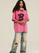 Frau in rosa Oversize-T-Shirt mit großem "B"-Logo, zerrissene Jeans, goldene Kopfhörer; Streetwear, urbaner Stil, modische Freizeitkleidung.