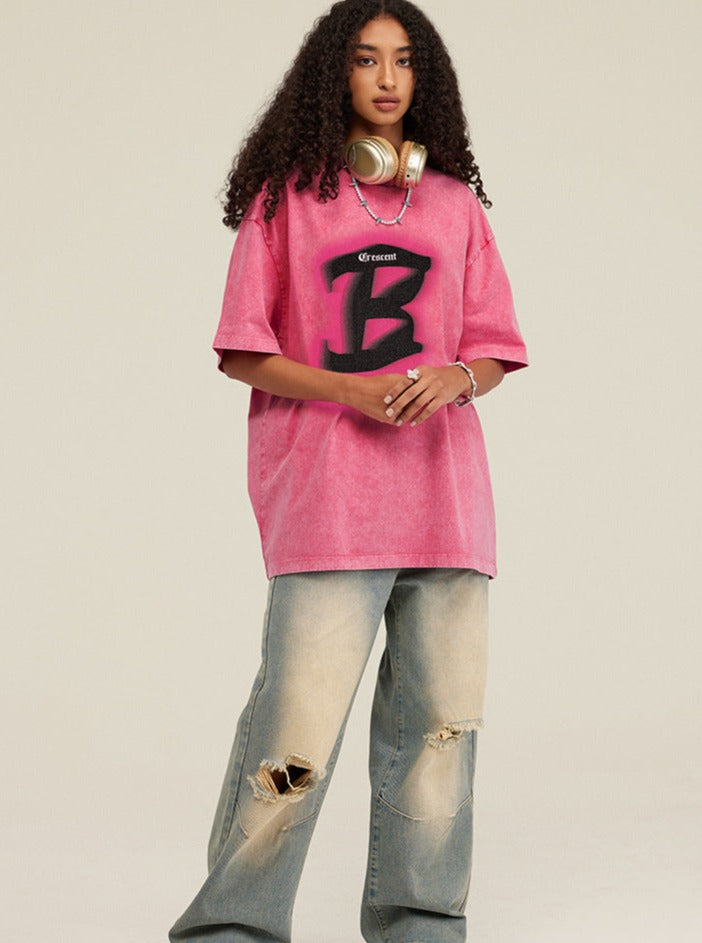 Frau in rosa Oversize-T-Shirt mit großem "B"-Logo, zerrissene Jeans, goldene Kopfhörer; Streetwear, urbaner Stil, modische Freizeitkleidung.
