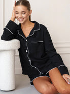 Frau in schwarzem Pyjama mit weißen Paspeln, sitzt lächelnd auf einem weißen Hocker. Bequeme Nachtwäsche, stilvoll und modern.
