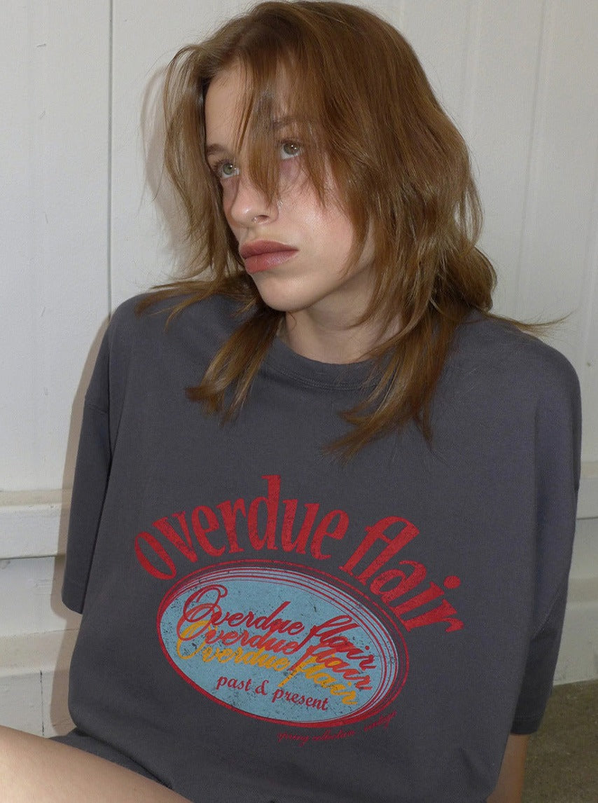 Graues T-Shirt mit rotem "overdue flair"-Aufdruck, getragen von einer Person mit braunem Haar. Modetrend, lässiger Stil, Streetwear.