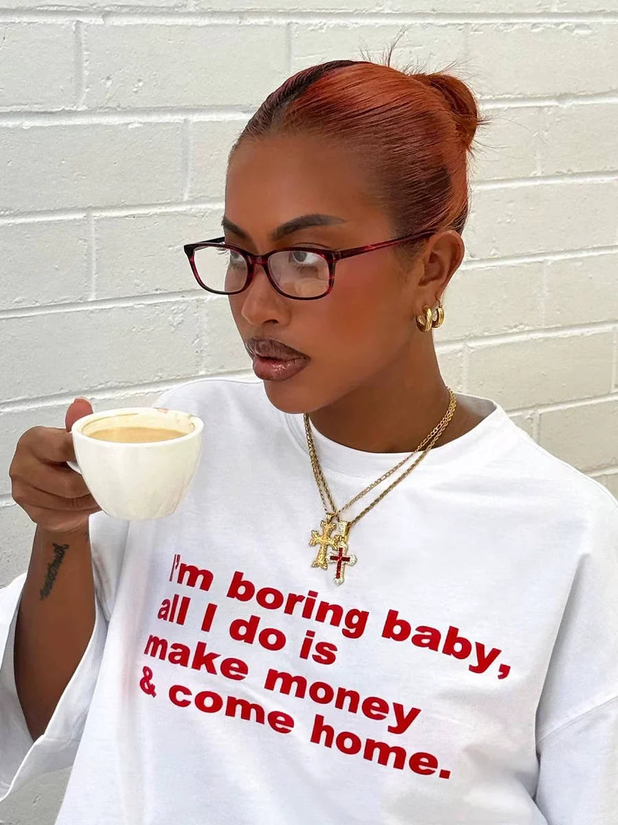 Frau mit Brille trinkt Kaffee, trägt weißes T-Shirt mit rotem Text, goldene Halsketten. Mode, Lifestyle, urbaner Stil, Kaffeegenuss.