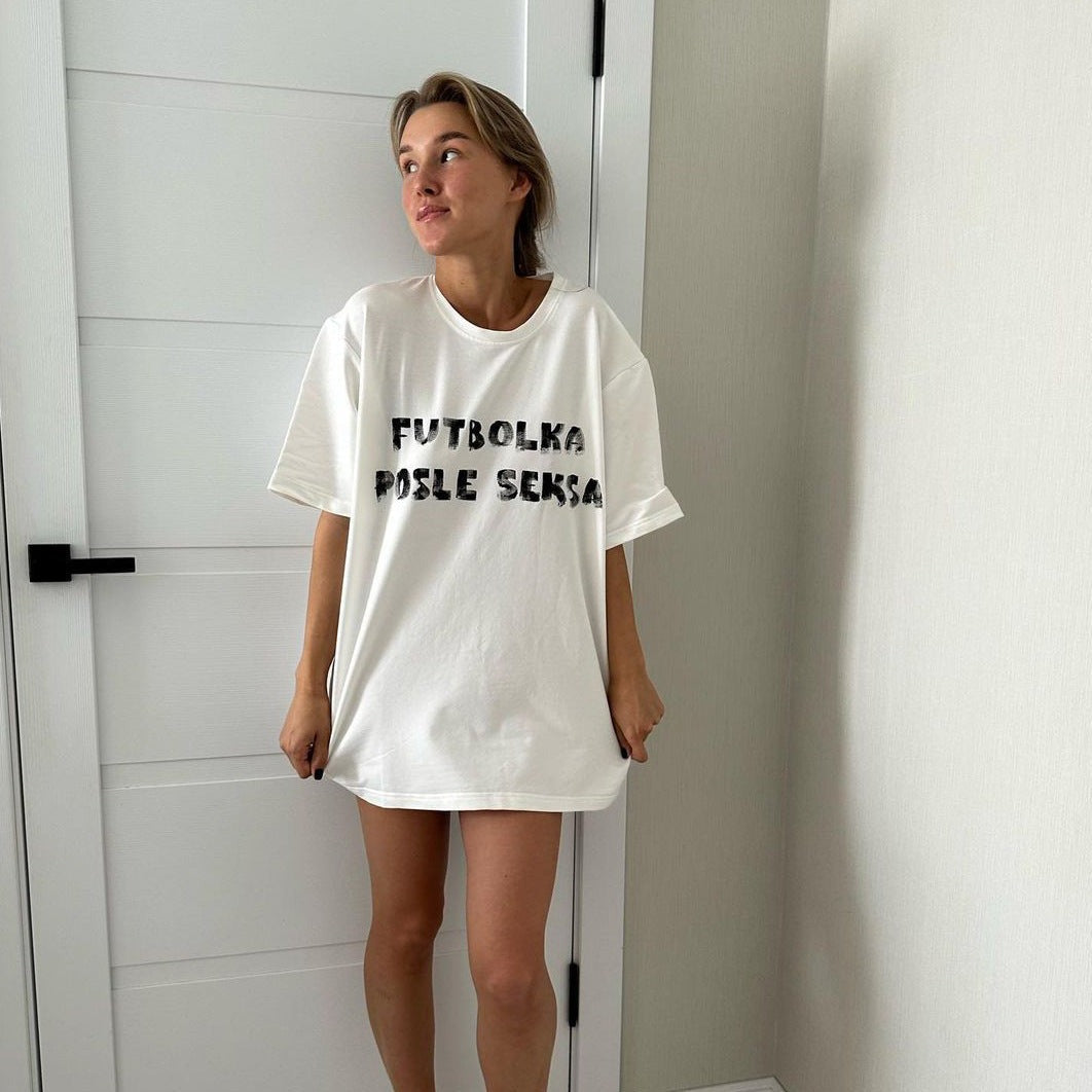 Frau trägt weißes T-Shirt mit schwarzem Aufdruck "Futbolka Posle Seksa", steht vor weißer Tür. Modetrend, lässiger Stil, Oversize-Shirt.
