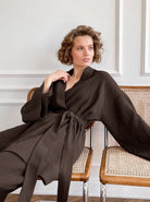 Frau in braunem Kimono aus Baumwolle sitzt auf einem Stuhl. Elegante Damenmode, bequeme Freizeitkleidung, stilvolle Homewear.
