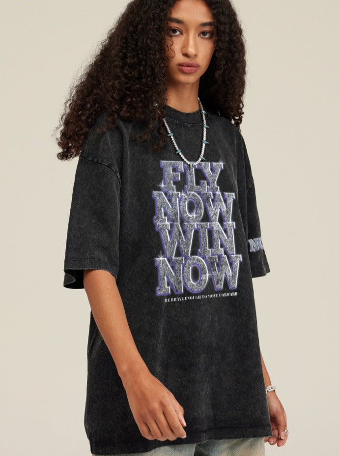 Schwarzes Oversize-T-Shirt mit "Fly Now Win Now"-Aufdruck, getragen von einer Person mit lockigem Haar. Trendige Streetwear-Mode für Jugendliche.