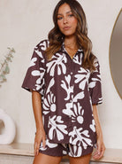Frau in braun-weißem, geblümtem Pyjama-Set aus Satin, steht vor neutralem Hintergrund. Modetrend, Nachtwäsche, Damenmode, bequem, stilvoll.