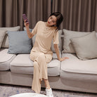 Frau in beigem Lounge-Set auf grauem Sofa, Smartphone in der Hand, moderne Wohnzimmereinrichtung, gemütliche Atmosphäre, stilvolle Inneneinrichtung.