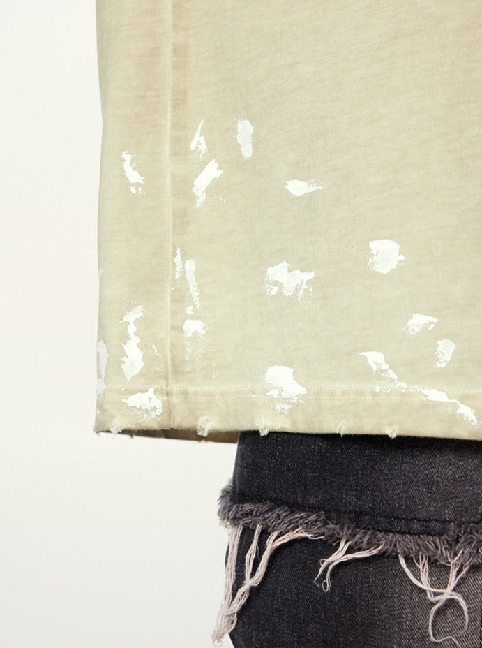 Beige T-Shirt mit weißem Farbklecks-Design, kombiniert mit zerrissenen schwarzen Jeans. Trendige Streetwear-Mode für lässigen Look.