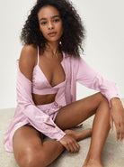 Frau in rosa Satin-Pyjama-Set, bestehend aus Hemd und Shorts, sitzt auf Teppich. Elegante Nachtwäsche, bequem und stilvoll.