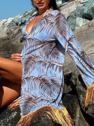 Frau in blauem Strandkleid mit Palmenmuster und Fransen sitzt auf Felsen. Sommermode, Strandkleidung, tropisches Design, Urlaubsoutfit.