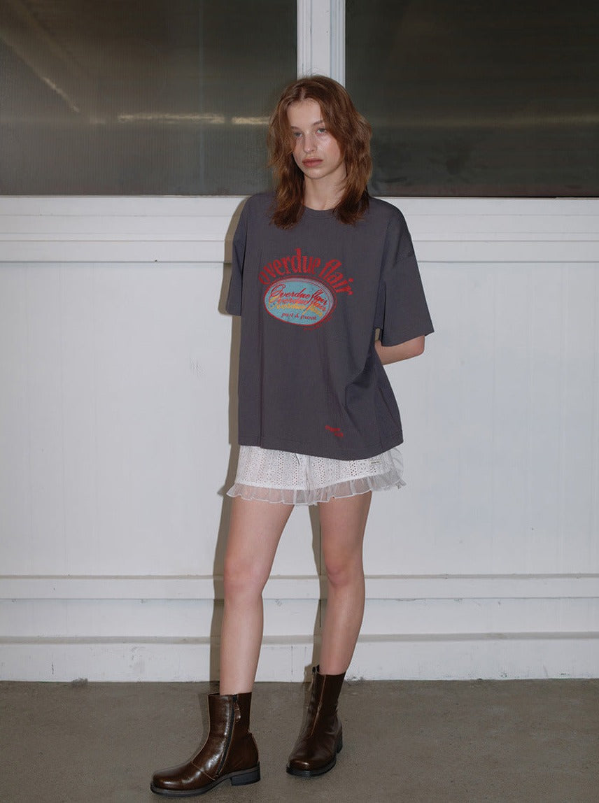 Frau trägt graues T-Shirt mit "Everyday Wear"-Aufdruck, kombiniert mit weißem Rock und braunen Stiefeln. Modetrend, lässiger Stil, urbaner Look.