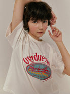 Junge Frau in weißem T-Shirt mit rotem "Overdue" Aufdruck, lässige Pose, modischer Look, trendige Damenmode, Streetwear, stilvolles Design.