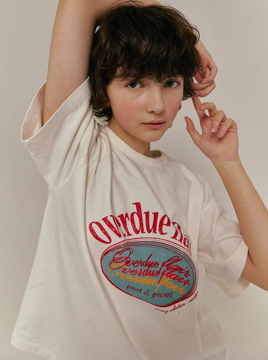 Junge Frau in weißem T-Shirt mit rotem "Overdue" Aufdruck, lässige Pose, modischer Look, trendige Damenmode, Streetwear, stilvolles Design.