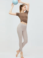 Frau in beiger Leggings und braunem T-Shirt hält blauen Fitnessball. Sportbekleidung, Yoga, Fitnessmode, athletische Kleidung, Workout-Outfit.