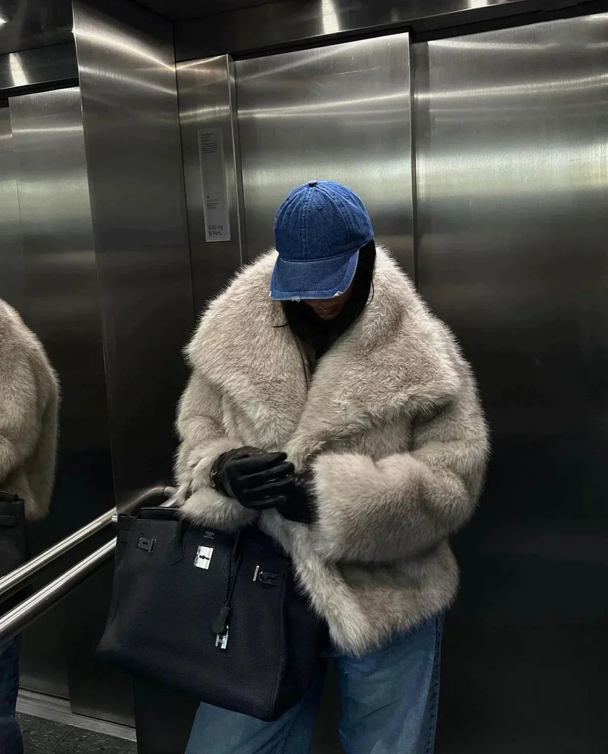 Person in einem Aufzug trägt eine luxuriöse, flauschige Pelzjacke, blaue Jeans und eine blaue Kappe, hält eine schwarze Designer-Handtasche.
