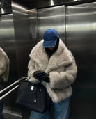 Person in einem Aufzug trägt eine luxuriöse, flauschige Pelzjacke, blaue Jeans und eine blaue Kappe, hält eine schwarze Designer-Handtasche.