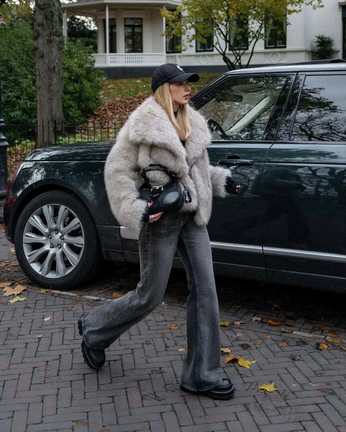Frau in grauem Kunstpelzmantel und Jeans, trägt schwarze Mütze und Kamera, geht neben grünem SUV auf gepflasterter Straße im Herbst.