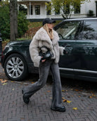 Frau in grauem Kunstpelzmantel und Jeans, trägt schwarze Mütze und Kamera, geht neben grünem SUV auf gepflasterter Straße im Herbst.